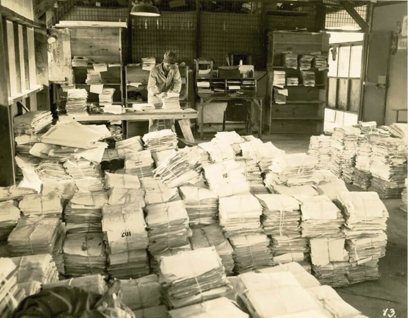 A person sits at a desk surrounded by large piles of stacked papers in a dimly lit, cluttered office space. Shelves and a cabinet are visible in the background. The image conveys a sense of overwhelming paperwork or archival work.